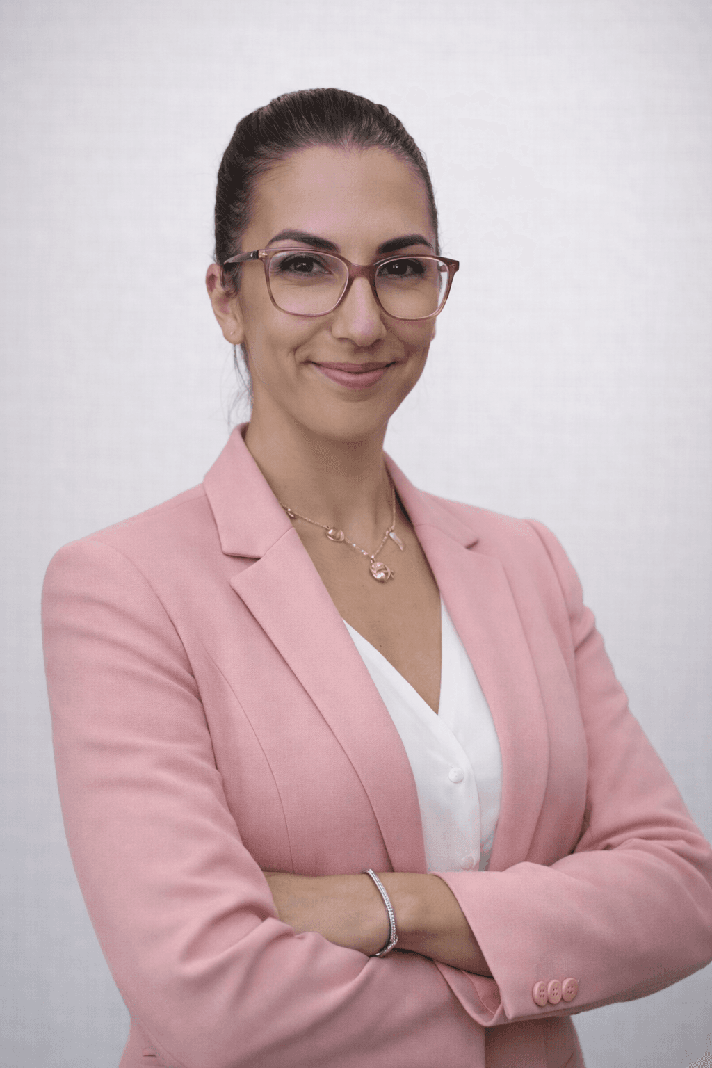 Portrait professionnel d'une femme souriante à lunettes, vêtue d'un blazer rose, les bras croisés.