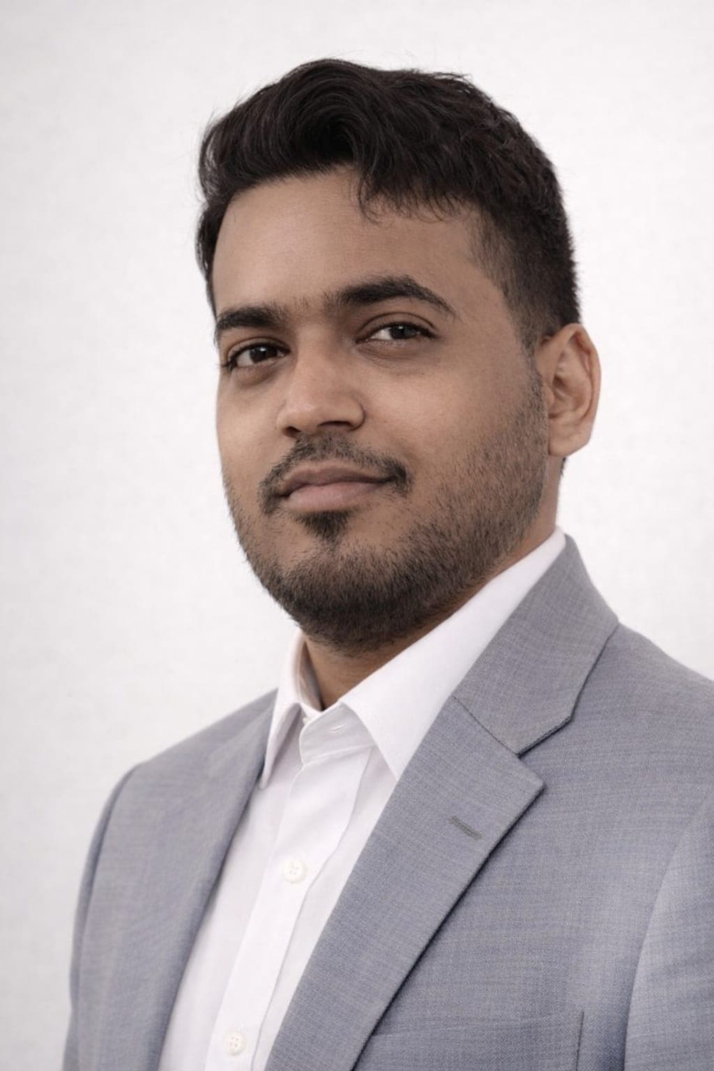 Homme barbu en costume gris et chemise blanche posant devant un fond blanc uni.